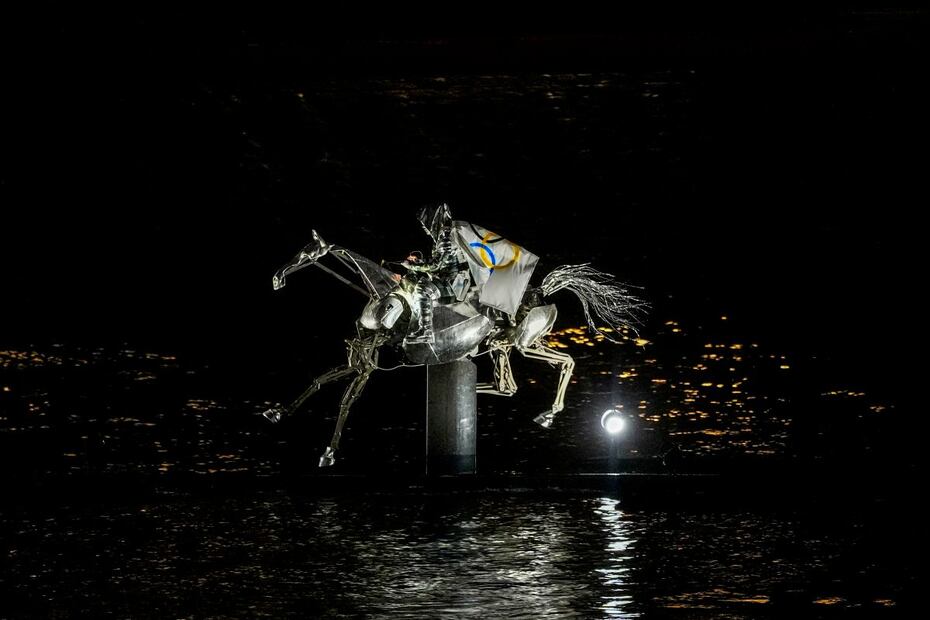 La doncella en armadura, con la bandera olímpica sobre un caballo mecánico / Foto: AP
